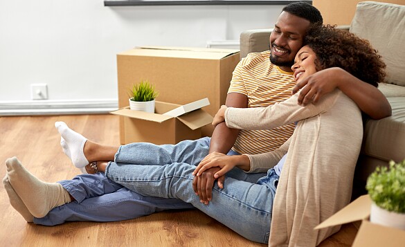 Ein junges Paar sitzt vor dem Sofa und umarmt sich. Im Hintergrund sind Umzugskartons zu sehen.