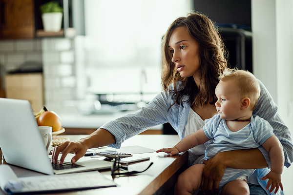 Eine Frau sitzt mit einem Baby zu Hause vor einem Laptop.