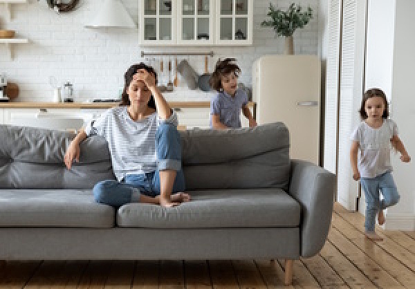 Erschöpfte junge Mutter auf Sofa, um sie herum rennen zwei Kinder