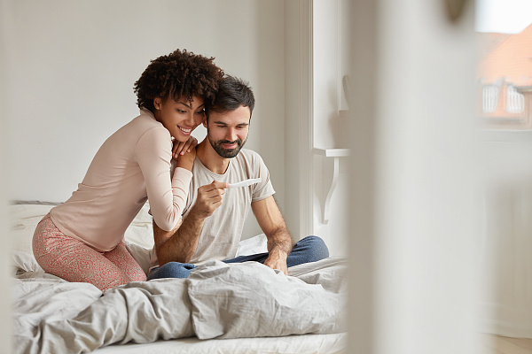 Eine Frau und ein Mann sitzen auf einem Bett. Der Mann hat einen Schwangerschaftstest in seiner Hand.