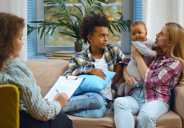 Eine Frau mit Klemmbrett sitzt vor einer dreiköpfigen Familie (Mutter, Sohn, Baby).